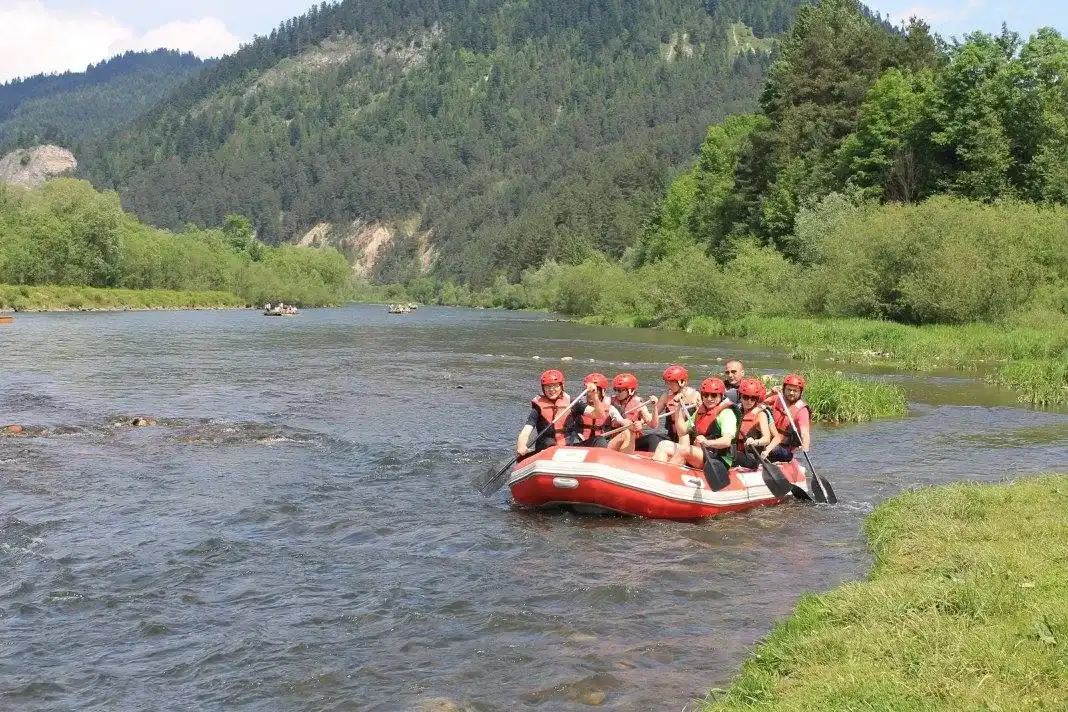 Grupa osób siedzi w pontonie na jeziorze, w kamizelkach ratunkowych i wiosłuje. Wokół nich są drzewa i trawa, pod czystym błękitnym niebem z górami w tle.