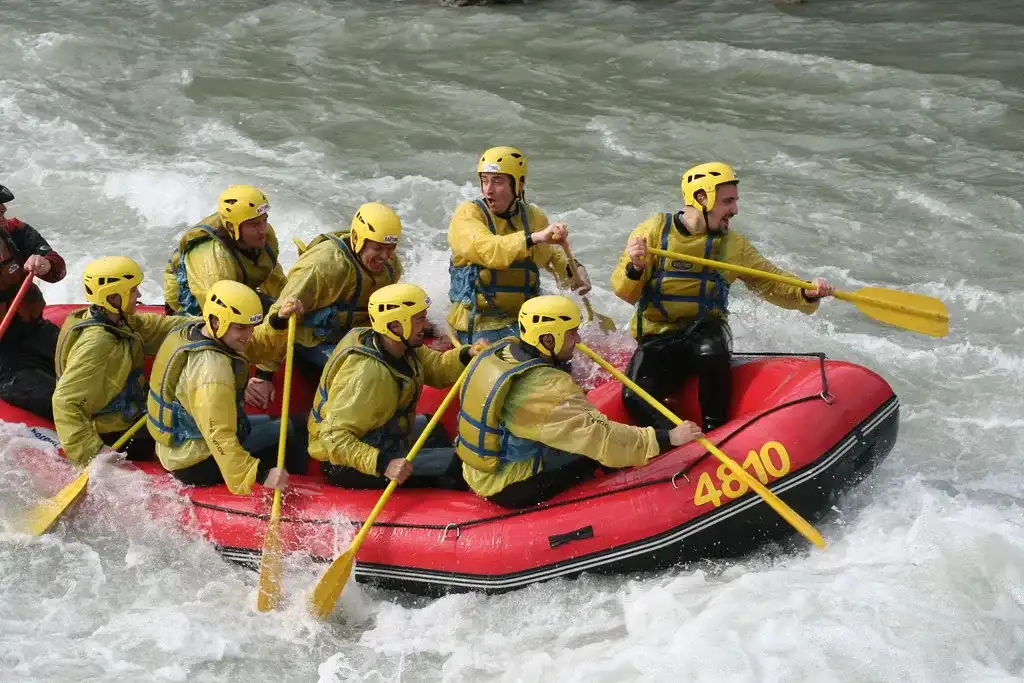 Grupa osób w kamizelkach ratunkowych płynie pontonem po rzece. Scena przedstawia dynamiczne warunki na świeżym powietrzu z pluskającą wodą i otaczającą przyrodą.