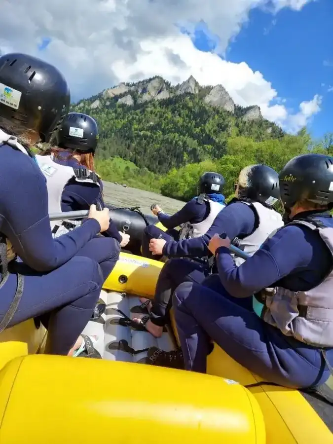 Grupa osób w pontonie na rzece otoczonej górami pod błękitnym niebem z chmurami.