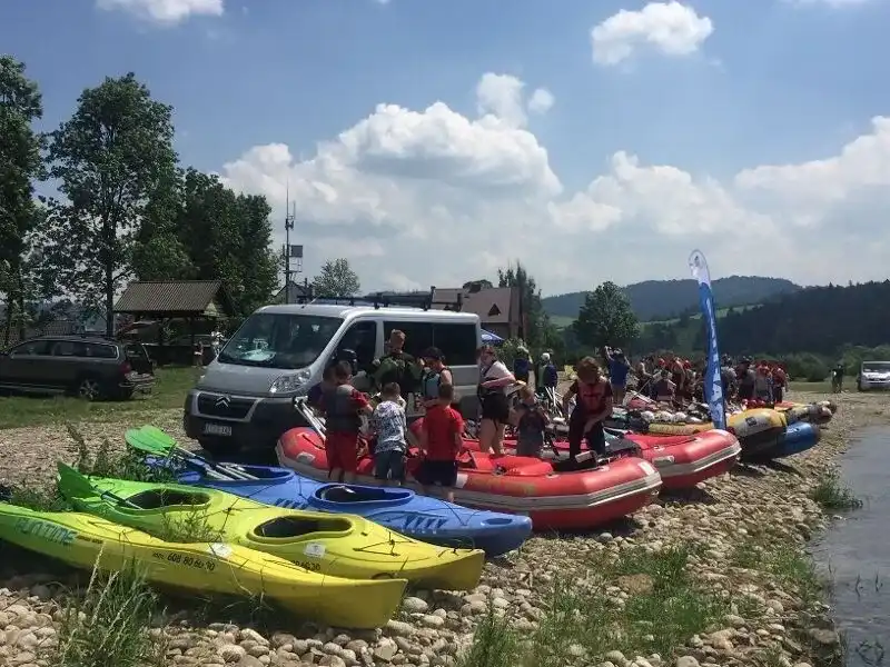 Grupa osób w kajakach zaparkowanych na skalistym brzegu, z drzewami w tle i częściowo zachmurzonym niebem. Mają na sobie kamizelki ratunkowe i wydają się cieszyć rekreacją na świeżym powietrzu.
