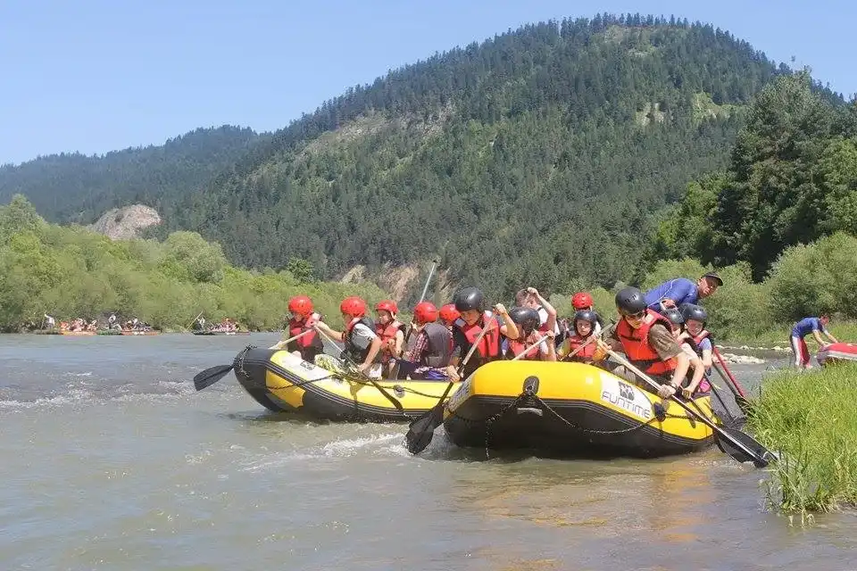 Grupa osób w kamizelkach ratunkowych siedzi w pontonie na rzece. Wspólnie wiosłują, z drzewami wzdłuż brzegu rzeki i czystym błękitnym niebem nad głową.