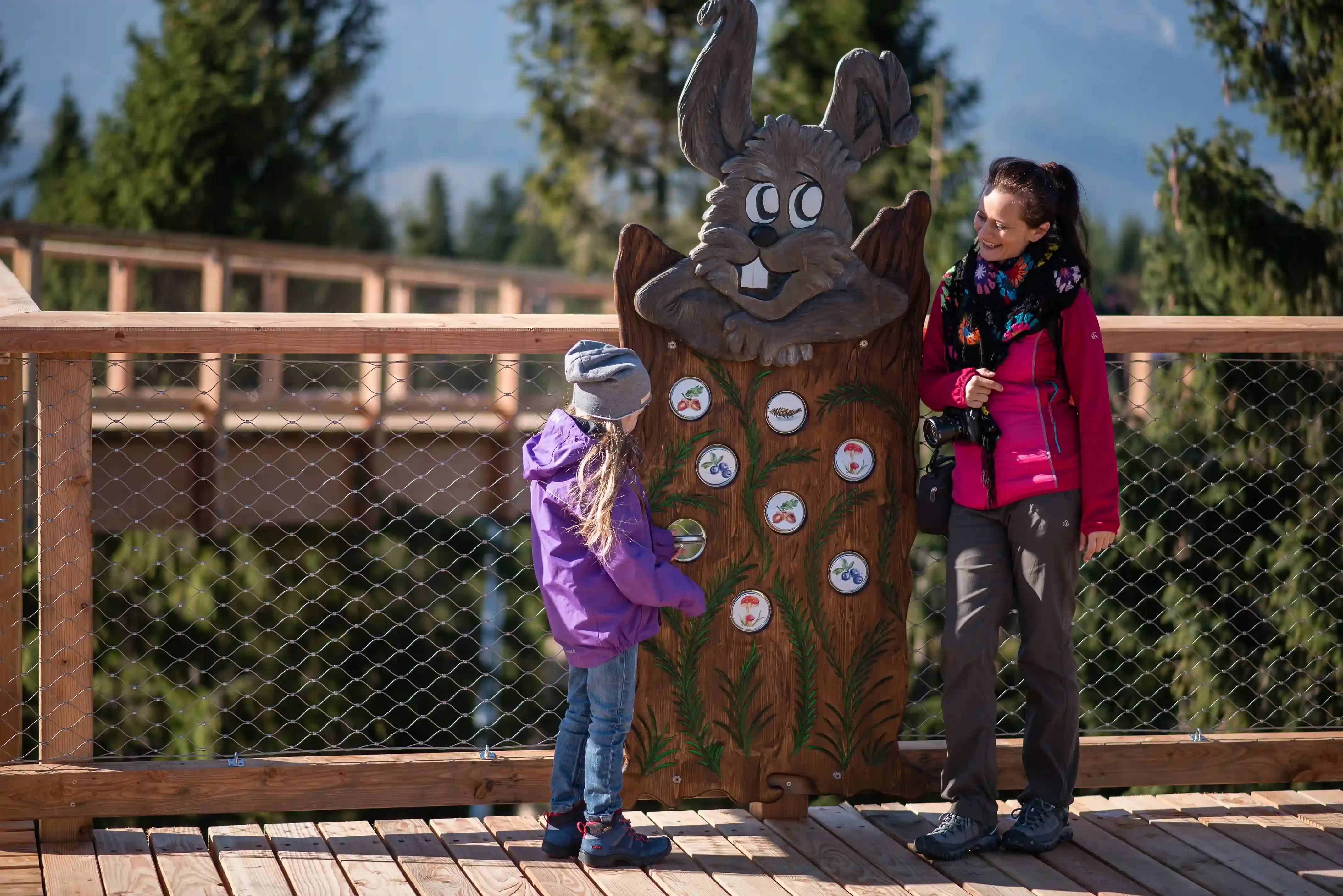 Kobieta i dziewczynka stojące przy drewnianej tablicy z królikiem, na zewnątrz pod błękitnym niebem, otoczone drzewami i płotem. Obie mają na sobie odzież odpowiednią do aktywności na świeżym powietrzu, widoczne jest także obuwie.