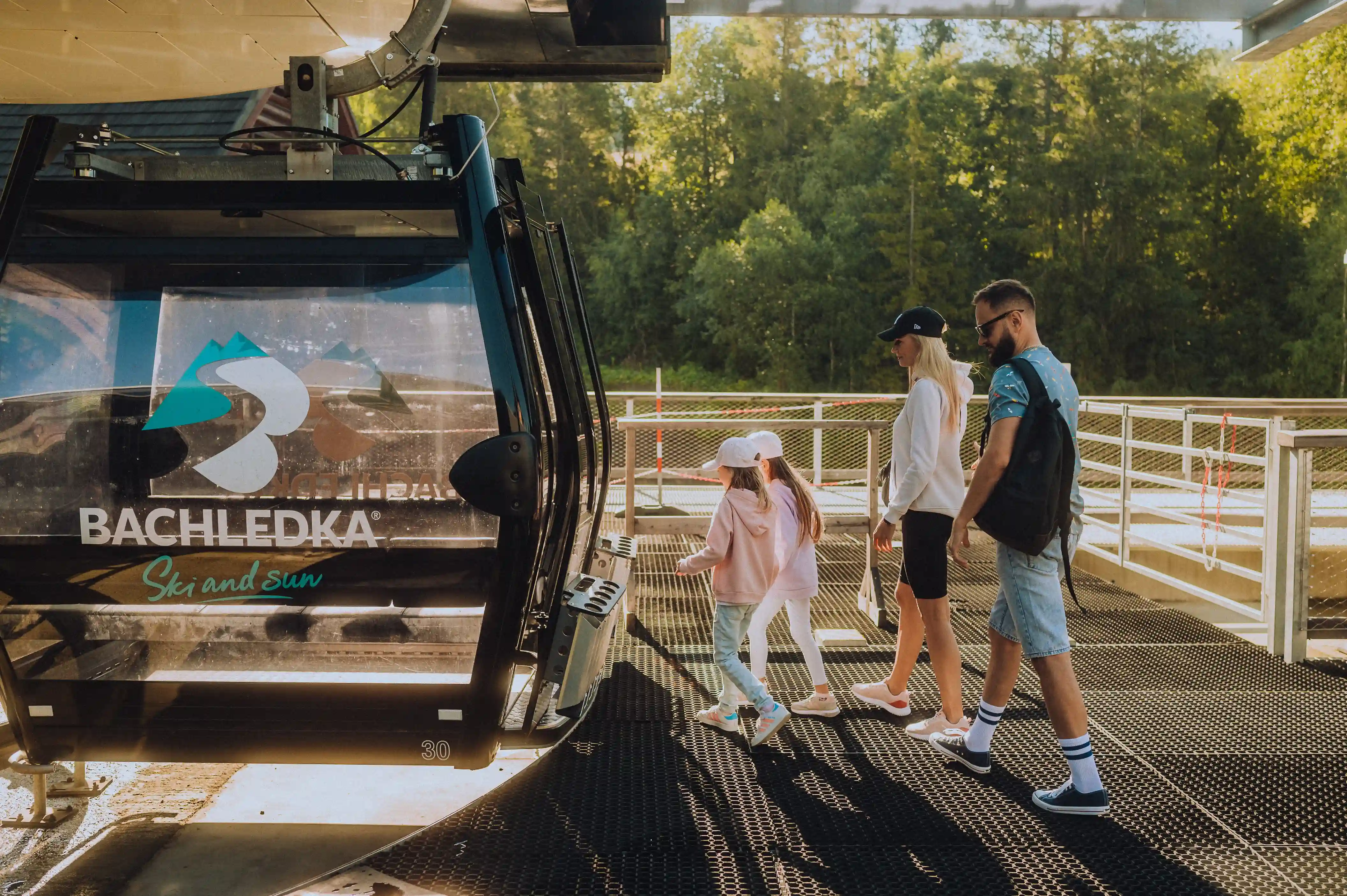 Grupa osób idących po platformie otoczonej drzewami, z ciężarówką i innymi pojazdami w tle. Jest słoneczny letni dzień, wszyscy są ubrani w casualowe ubrania i obuwie.