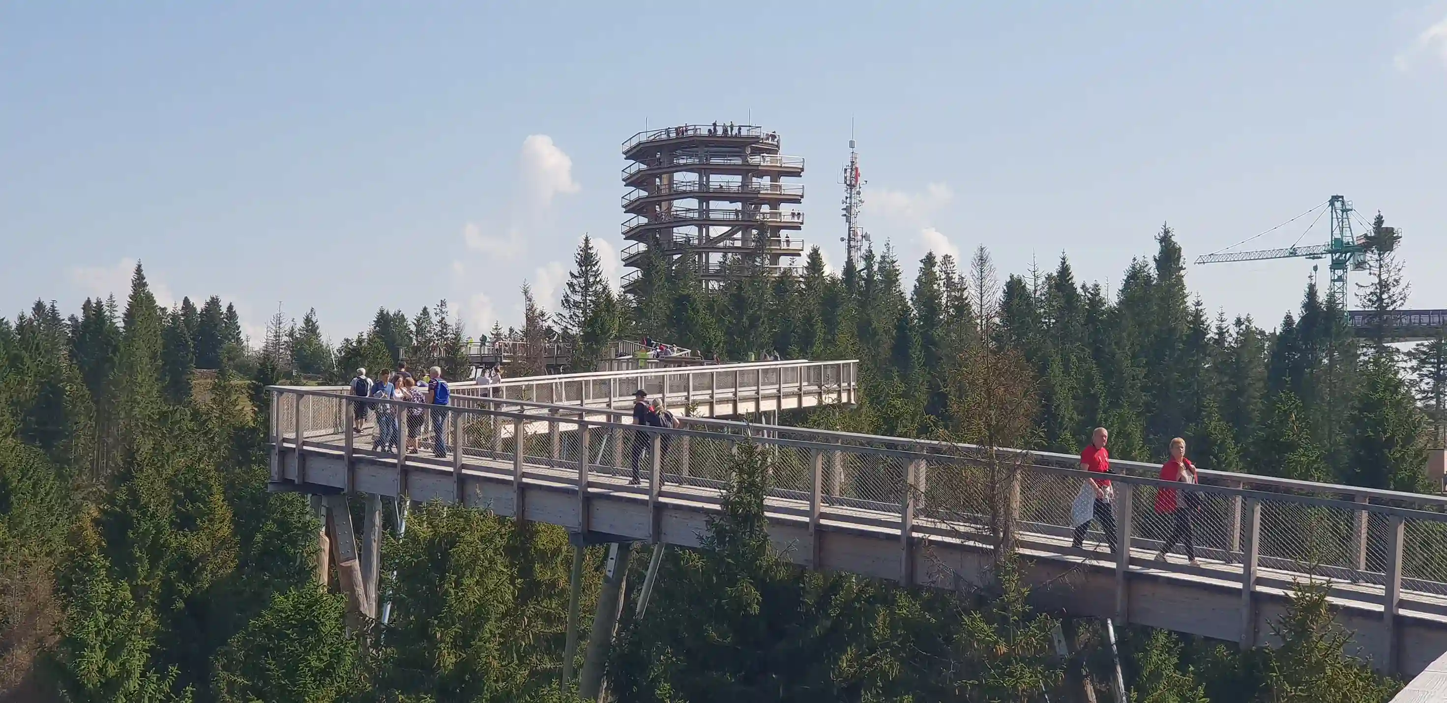Most, po którym przechodzi kilka osób, otoczony drzewami pod częściowo zachmurzonym niebem.