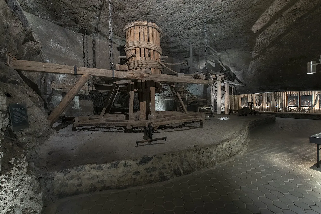 Drewniana konstrukcja znajduje się w jaskini, otoczona skalnymi ścianami i słabym oświetleniem. Grunt jest nierówny i naturalny, uzupełniając rustykalny wygląd drewnianej konstrukcji.