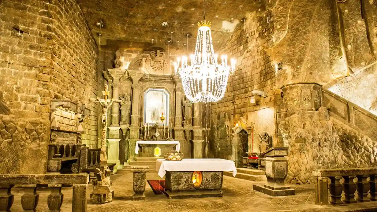 Dekoracyjny żyrandol wisi z sufitu pomieszczenia z kamiennymi ścianami i kamienną podłogą. Przestrzeń jest umeblowana stołem, tworząc atmosferę wnętrza przypominającą kościół lub historyczny budynek.