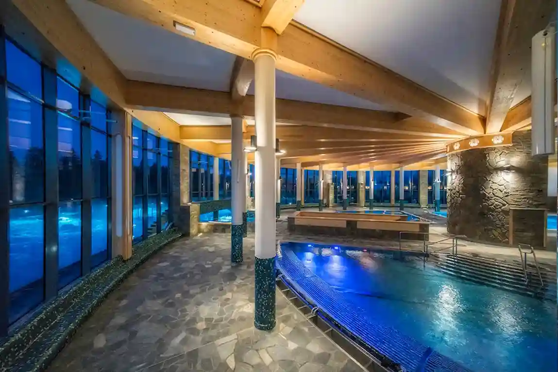 Przestronny kryty basen z dużą kamienną podłogą i dużym oknem, które wpuszcza naturalne światło. Basen jest otoczony ścianami i ma sufit nad głową, tworząc relaksującą atmosferę w środowisku centrum rekreacyjnego.