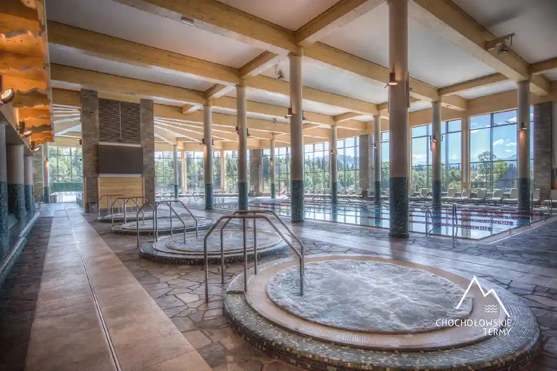 Przestronny kryty basen otoczony dużą kamienną podłogą, z oknami wpuszczającymi naturalne światło dzienne. Pomieszczenie ma wysokie sufity i ściany, które przyczyniają się do otwartej atmosfery.
