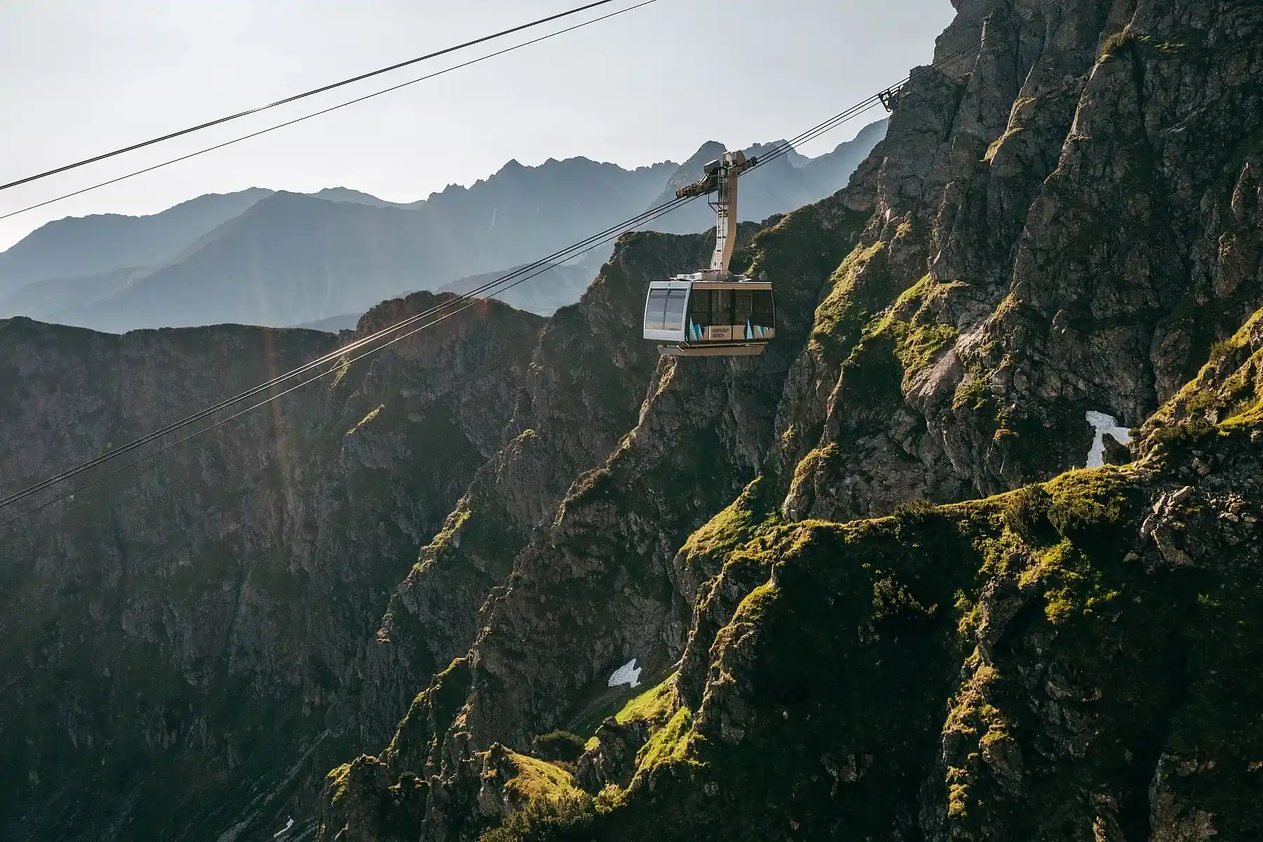 Kolejka linowa sunąca nad górskim krajobrazem z pagórkami, mgłą i klifami w tle pod czystym niebem.