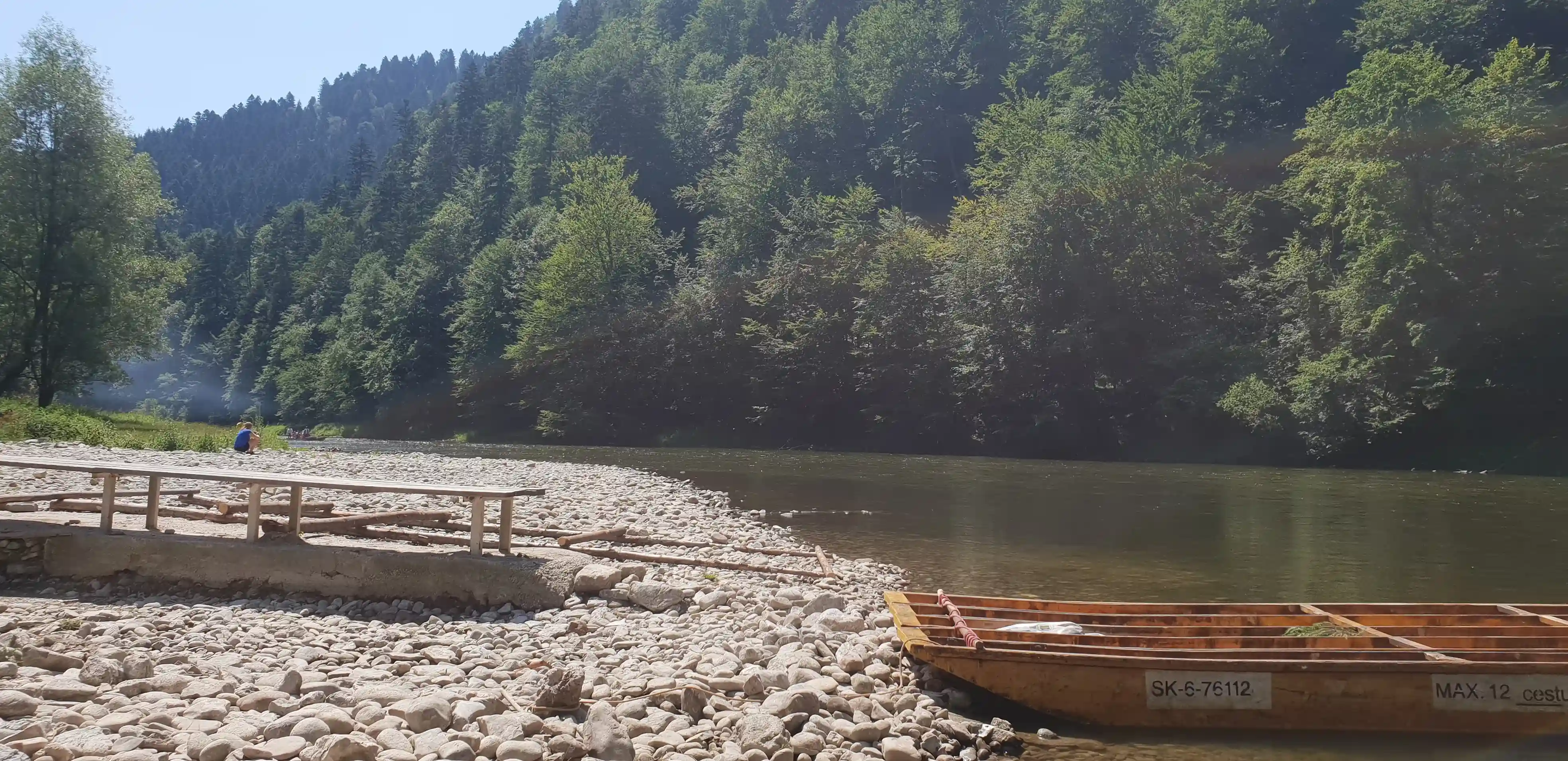 Łódź spoczywająca na skalistym brzegu obok rzeki, otoczona drzewami i górami pod bezchmurnym niebem.