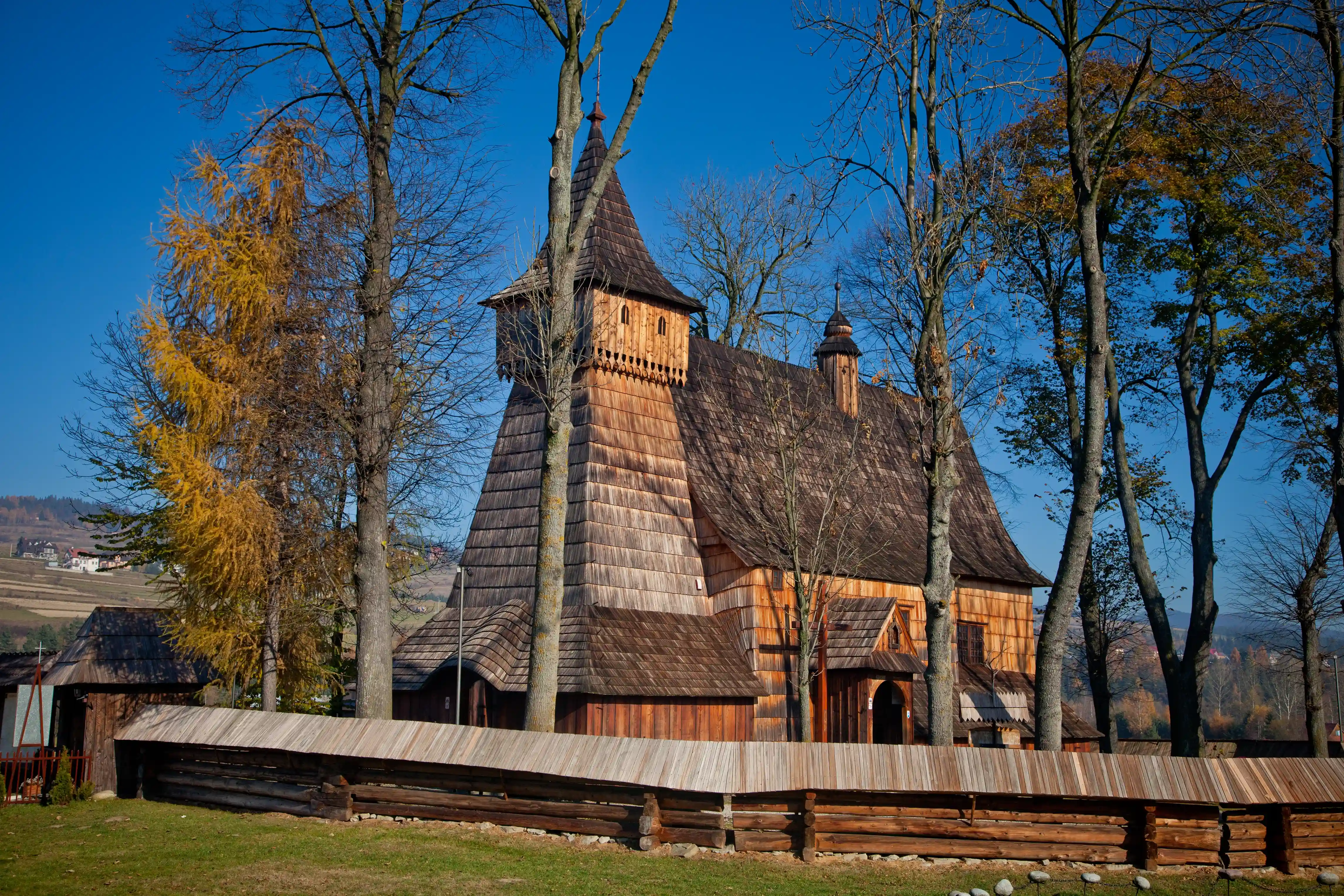 Drewniany budynek ze spiczastym dachem otoczony drzewami i trawą, pod bezchmurnym niebem w jesiennym krajobrazie.