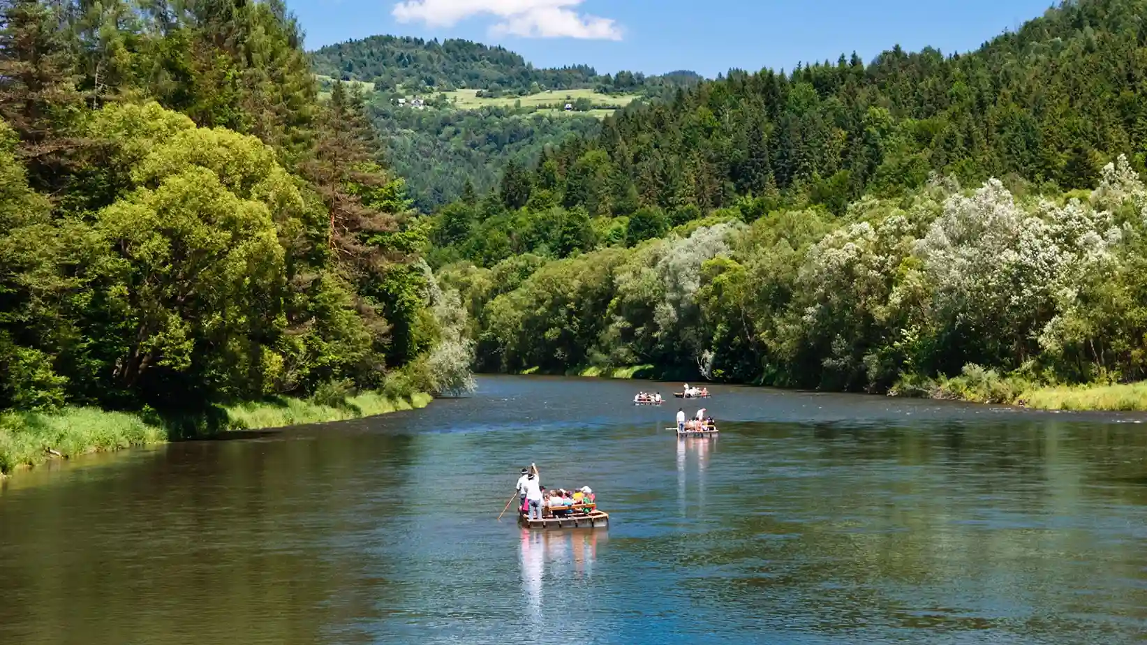 Grupa ludzi w łodzi na rzece otoczonej drzewami i górami pod pochmurnym niebem.