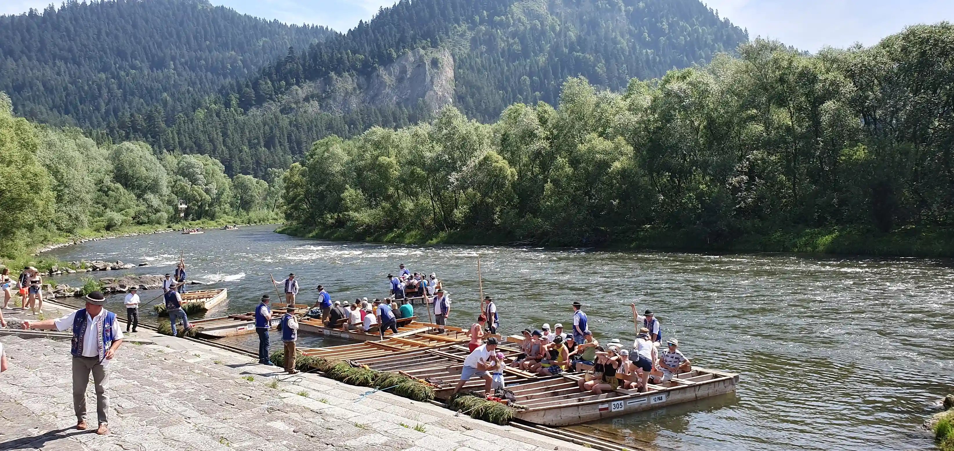 Grupa ludzi ciesząca się nawigowaniem tratwą na spokojnym zbiorniku wodnym otoczonym drzewami i górami pod bezchmurnym niebem.