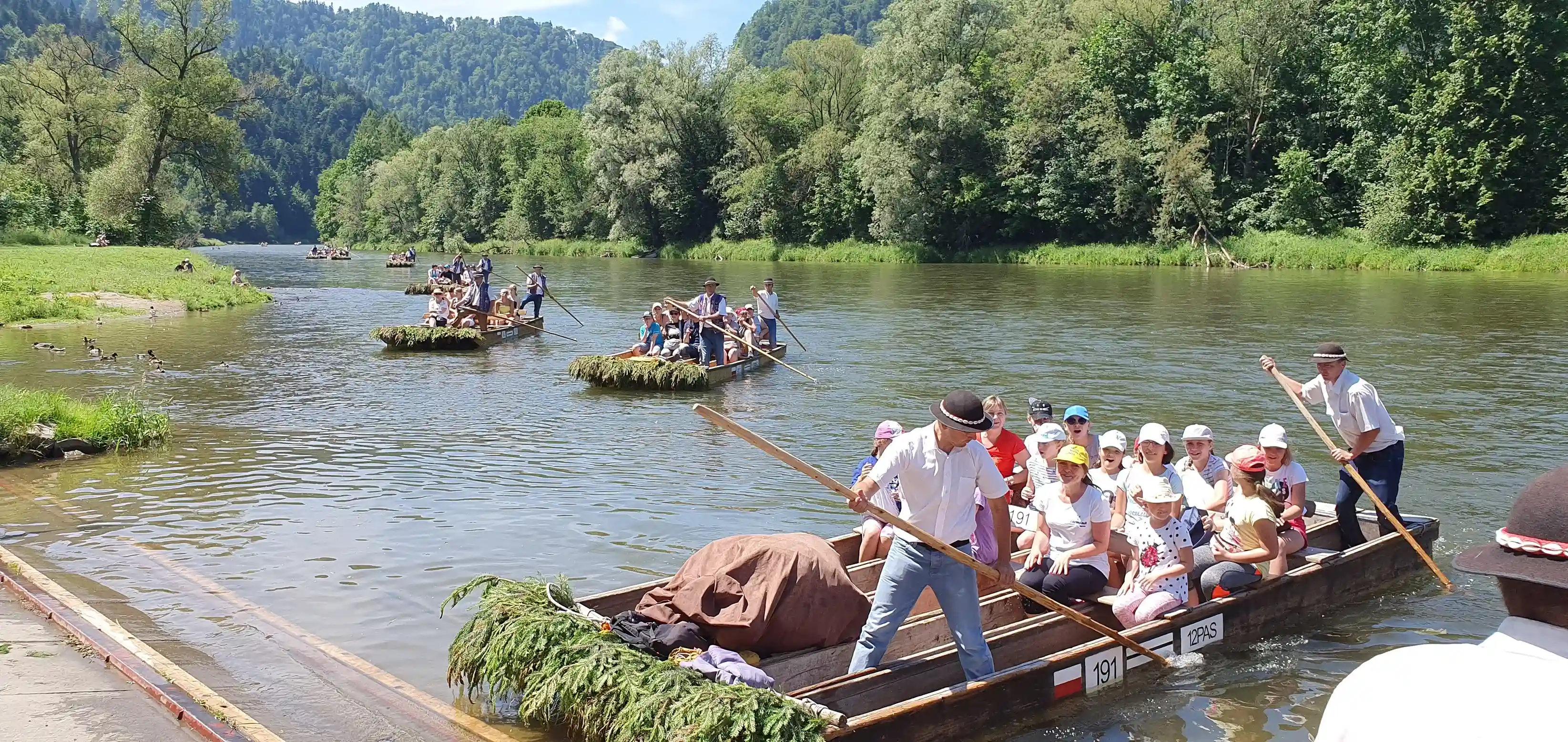 Grupa ludzi w łodzi wiosłujących w dół rzeki otoczonej drzewami i górami.