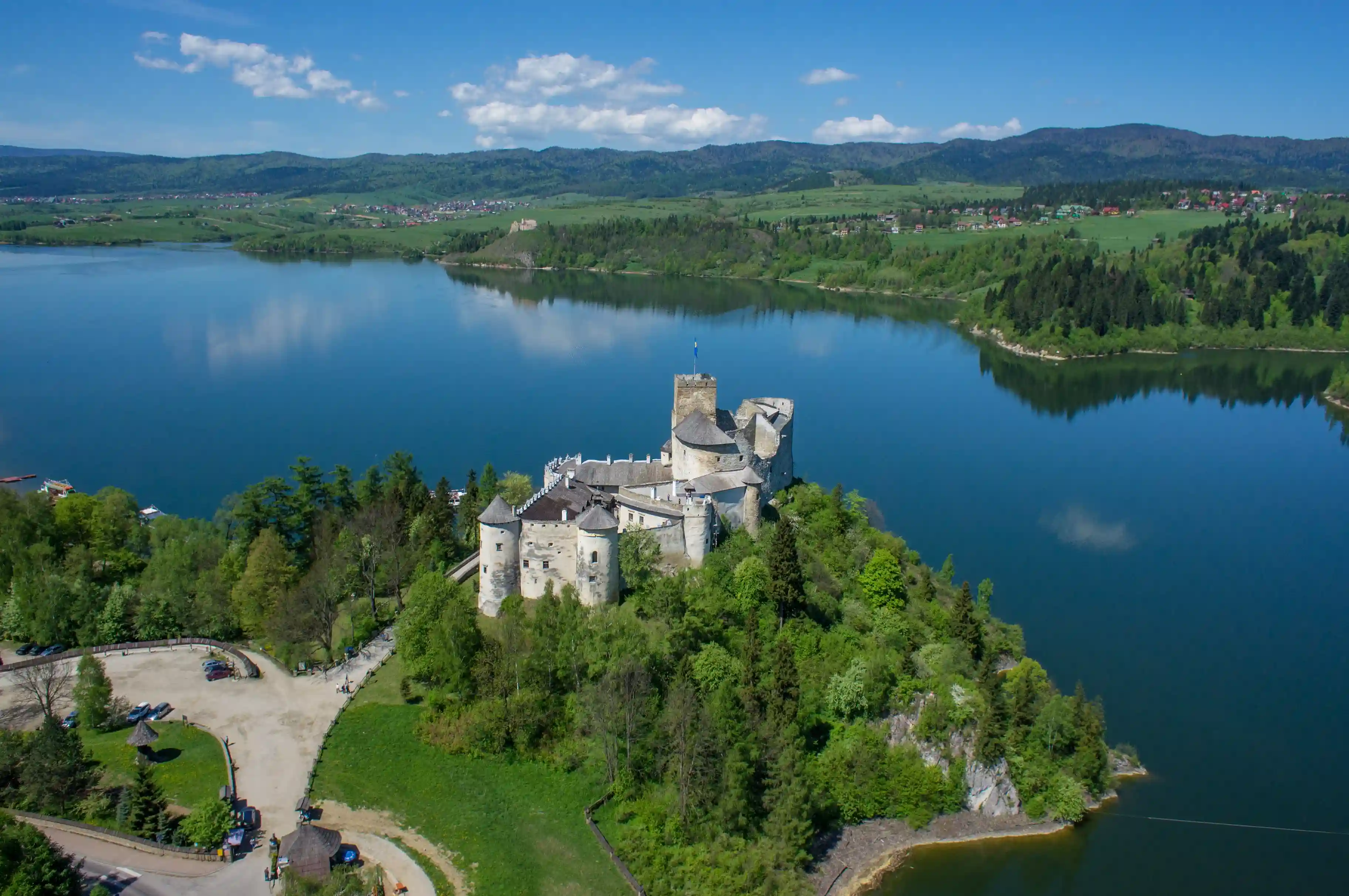 Zamek położony na wyspie, otoczony wodą z drzewami i bezchmurnym niebem w tle. Krajobraz obejmuje zbiornik wodny i góry, a także chmury nad głową. Zielona trawa porasta brzeg zbiornika wodnego.