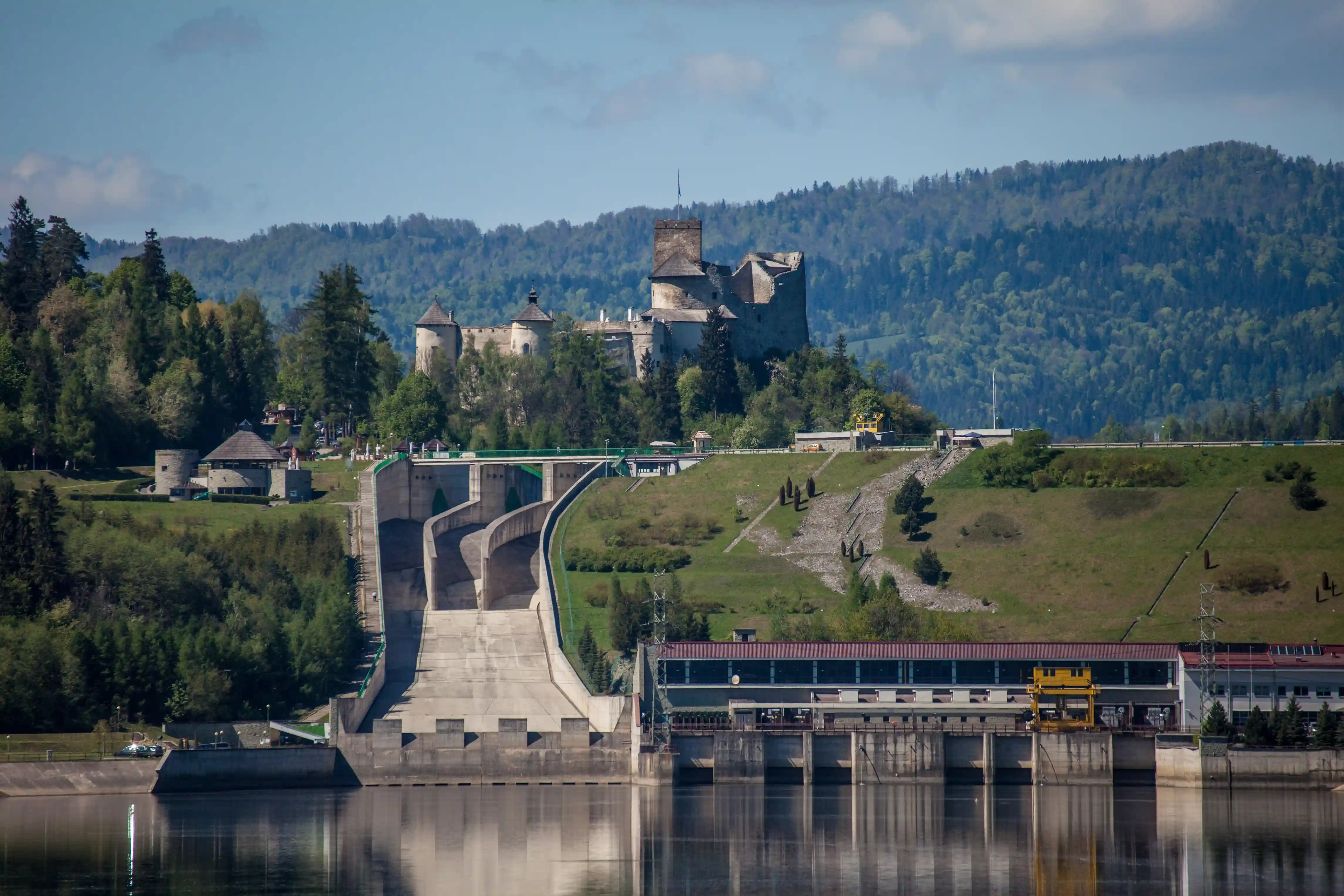 Zamek usytuowany na wzgórzu z widokiem na zbiornik wodny, otoczony drzewami i górami pod pochmurnym niebem. Most rozciąga się nad zbiornikiem, w pobliżu widoczny jest pociąg w krajobrazie.